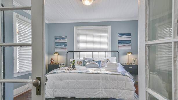 A bedroom with a bed, two bedside tables, a window with blinds, and wall art. - Cozy 2nd-Floor Suite near Beach, The Strand, Pier (Galveston)