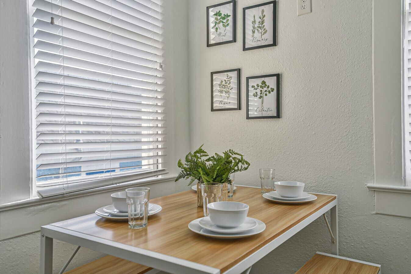 Un coin repas avec une table en bois, des assiettes blanches et des verres, un vase rempli de feuillage vert et, au mur, des illustrations botaniques encadrées.