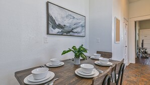 A dining area with a wooden table set for four, white plates, bowls, and cutlery. A potted plant is on the table, and a framed artwork hangs above. - 3 Room 2nd-Floor Suite near Beach, Pier, Strand (Galveston)