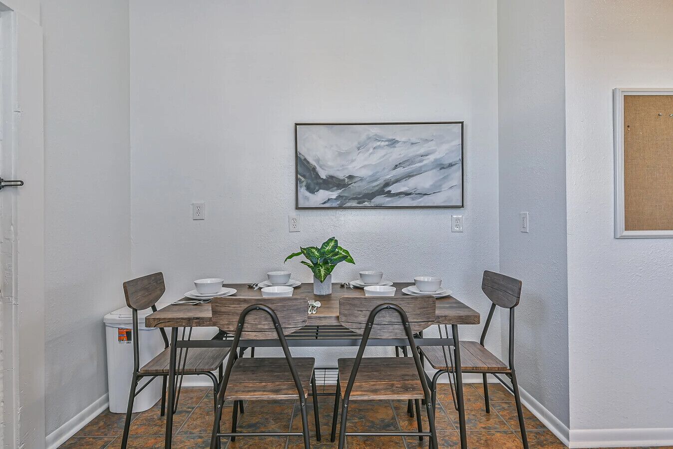 Un comedor con una mesa dispuesta para cuatro personas, una planta en maceta y un cuadro abstracto en la pared.