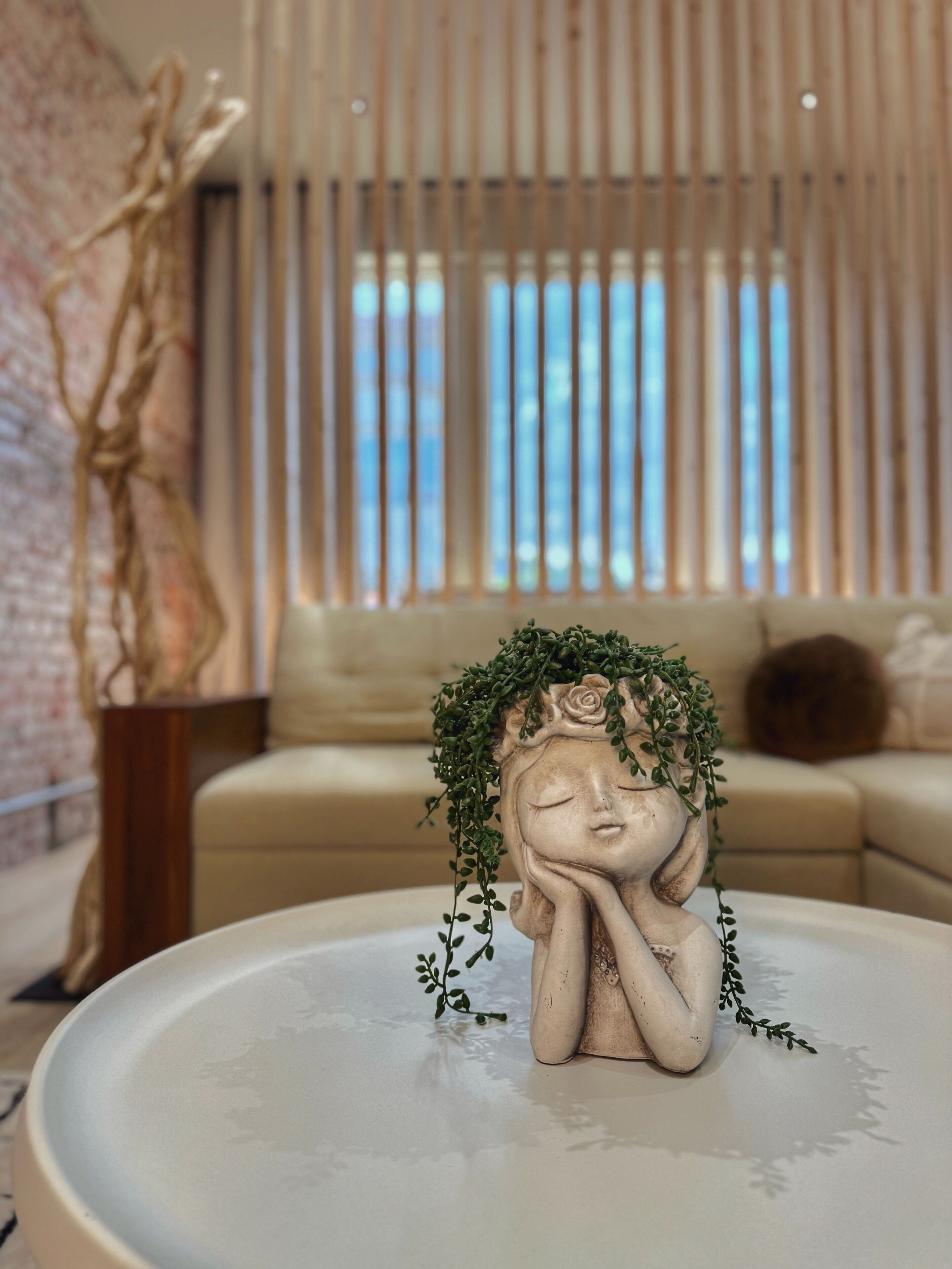 Une statue représentant une femme coiffée d’une couronne de verdure, posée sur une table blanche dans une pièce où se trouvent un canapé et une fenêtre équipée de stores verticaux.