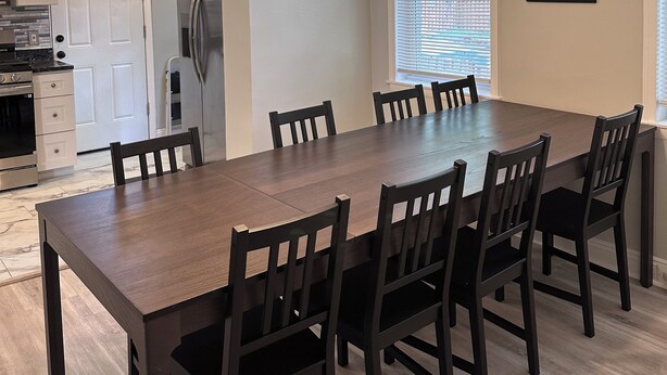 A dining area with a wooden table and chairs, a chandelier, a refrigerator, and a window with blinds.