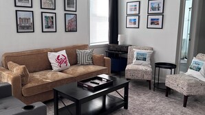 A living room with a brown sofa, a black coffee table, and a patterned armchair. There are framed pictures on the wall and a window with blinds.