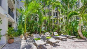 Une cour aménagée avec des fauteuils et des plantes en pot, entourée de grands palmiers et d’un immeuble à plusieurs étages.