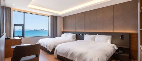 A modern hotel room with two beds, a desk, and a view of the sea.