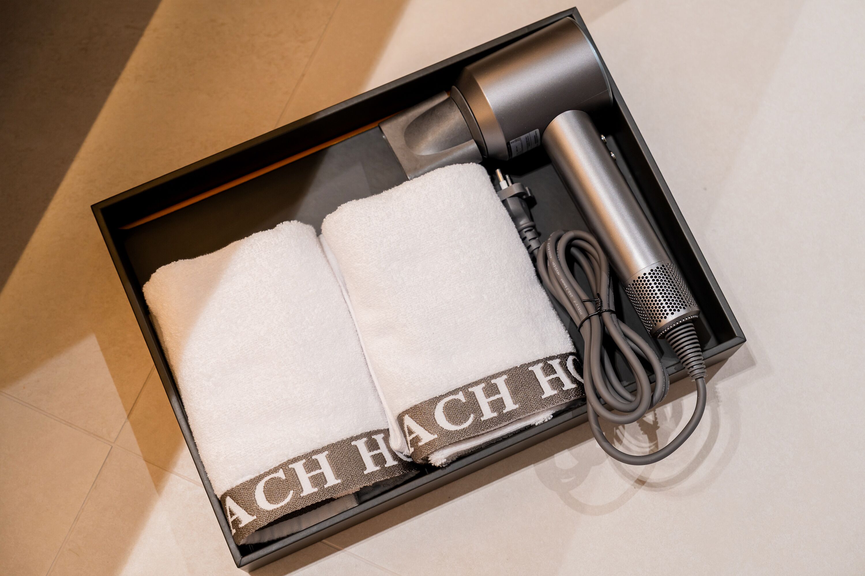 A black box containing a white towel with "ACH HACH HACH" text, a black hairdryer, and a black charging cable.