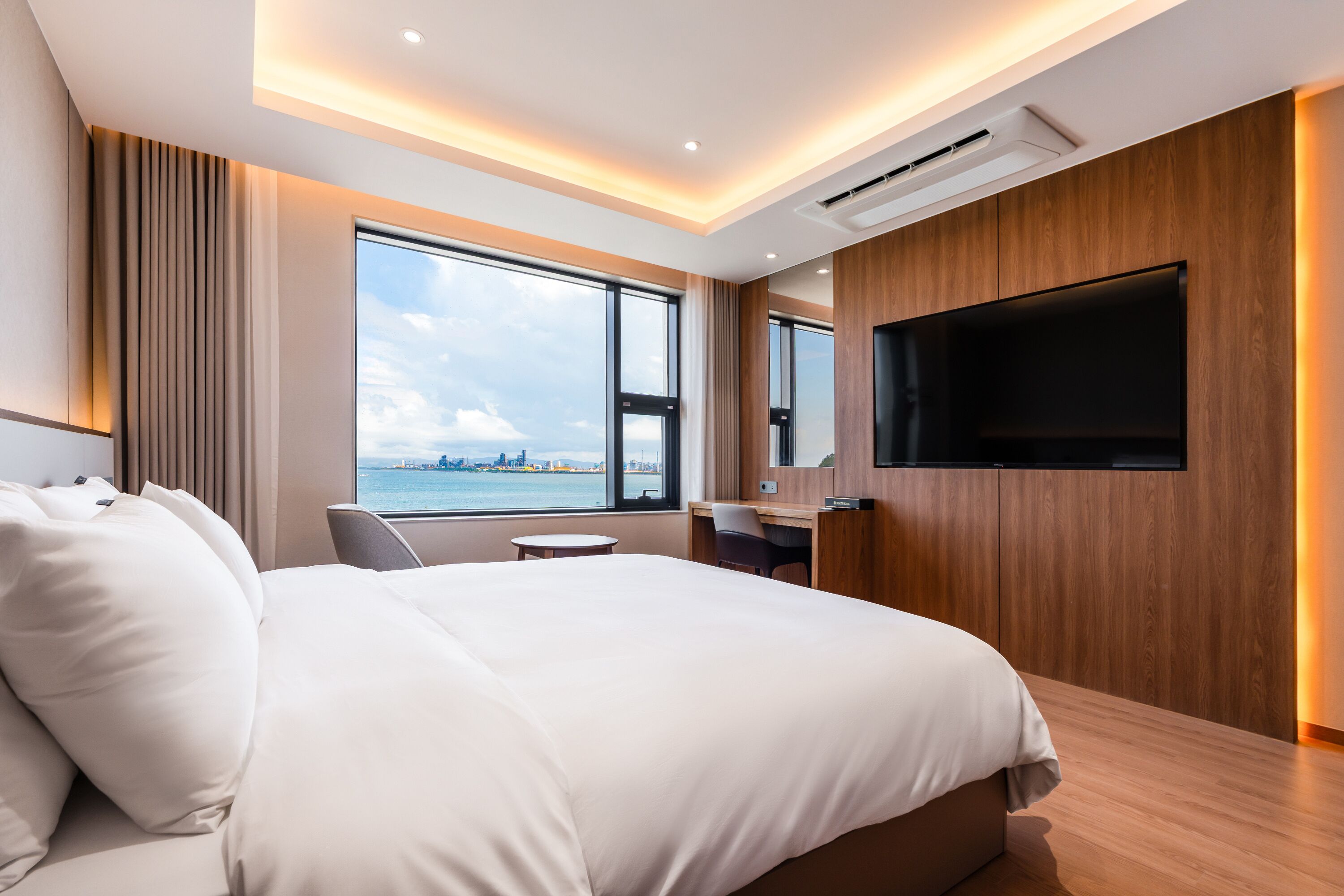 A modern hotel room with a large bed, a desk, a chair, a TV, and a window with a city view.