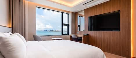 A modern hotel room with a large bed, a desk, a chair, a TV, and a window with a city view.