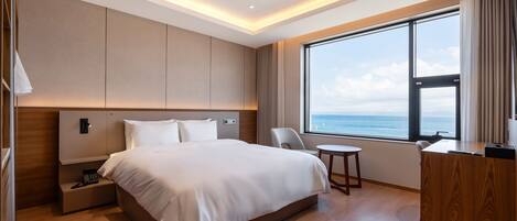 A modern hotel room with a large bed, a desk, a chair, and a view of the sea.
