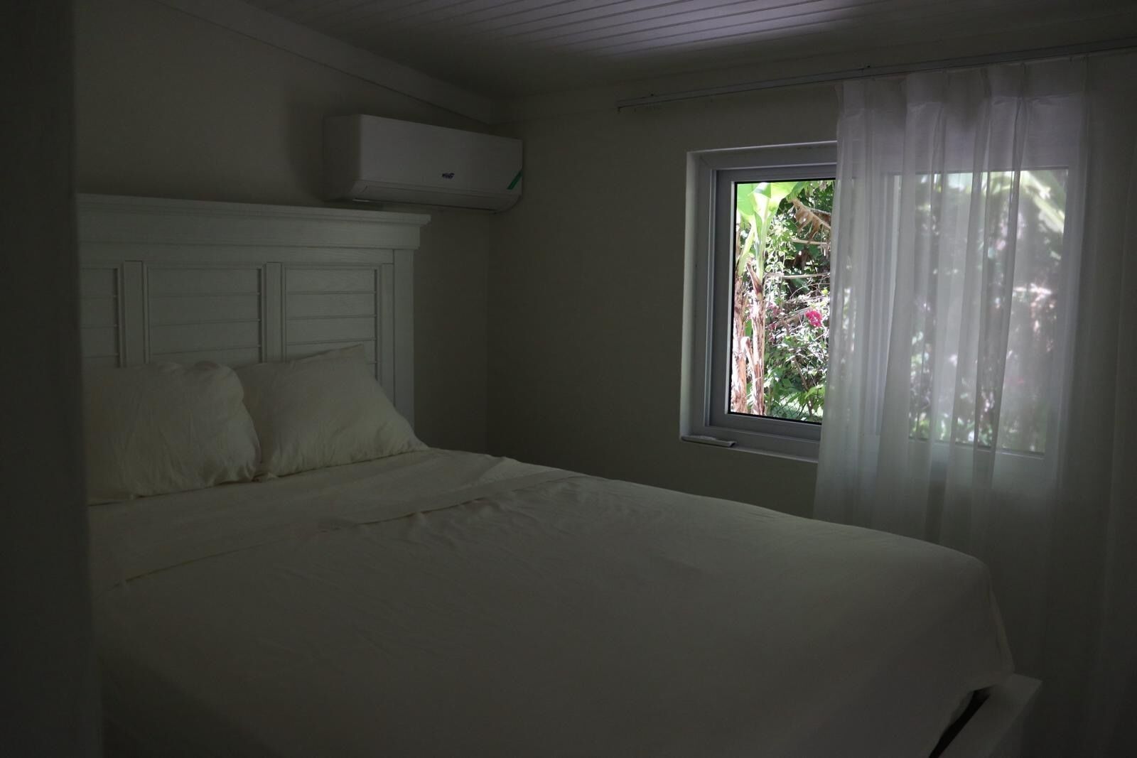 A bedroom with a bed, an air conditioner, a window with curtains, and a view of greenery outside.