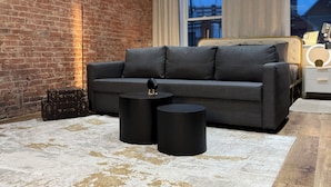 A modern living room with a brick wall, a grey sofa, two black side tables, and a white rug.
