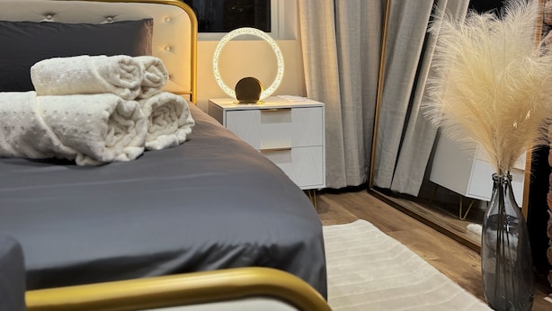 A modern bedroom with a large bed, a bedside table with a mirror, a vase with pampas grass, and a view of the city at night.