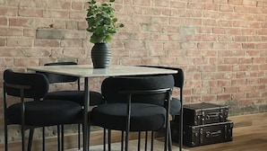 A dining area with a brick wall, a round table, four black chairs, a potted plant, and a suitcase on the floor.