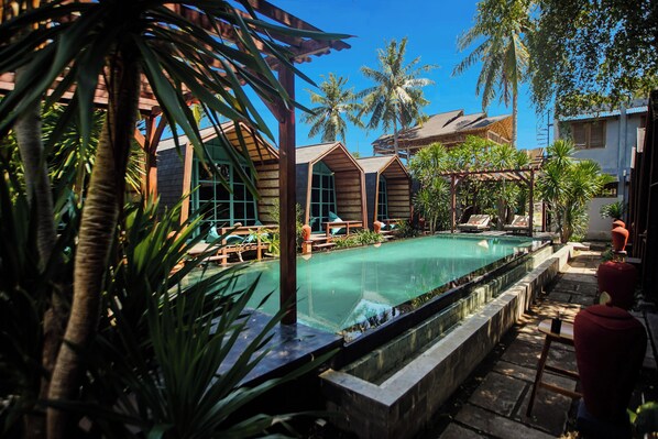 A pool area with lounge chairs, palm trees, and wooden buildings. - Waras Villa (Gili Trawangan)