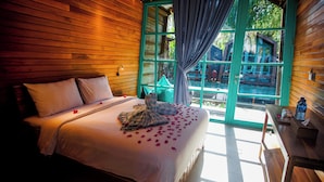 A wooden room with a bed, a window with curtains, and a table with a bottle and a glass. - Waras Villa (Gili Trawangan)