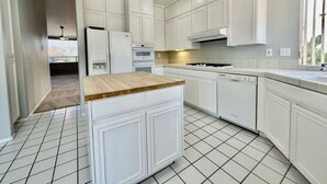 Une cuisine avec des meubles blancs, un îlot central doté d’un plan de travail en bois et un sol carrelé.