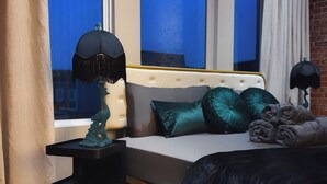 Un dormitorio moderno con una cama grande, una mesita de noche, una lámpara y vistas a la ciudad de noche.