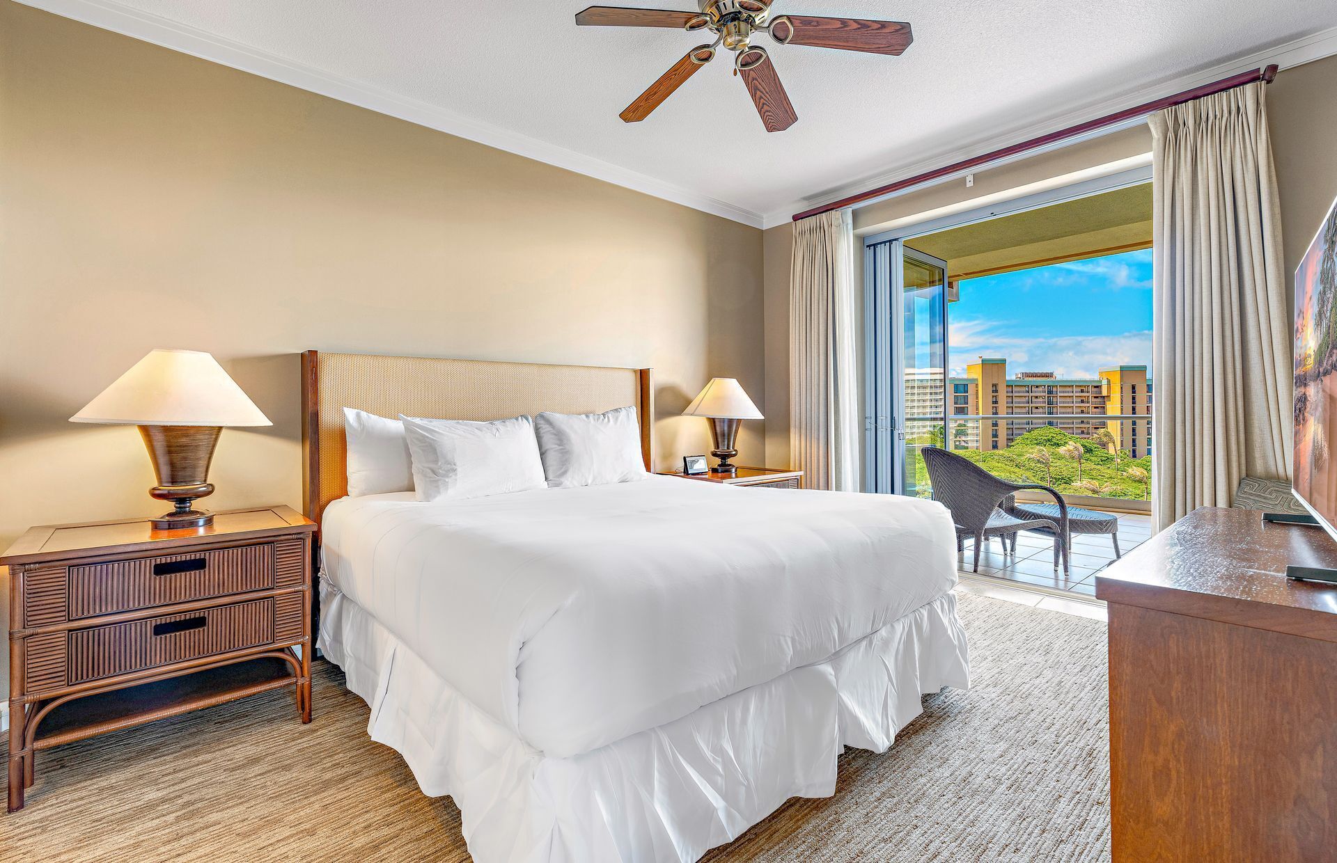 A hotel room with a large bed, two bedside lamps, a wooden nightstand, a desk with a chair, a ceiling fan, and a balcony with a view of buildings and greenery.