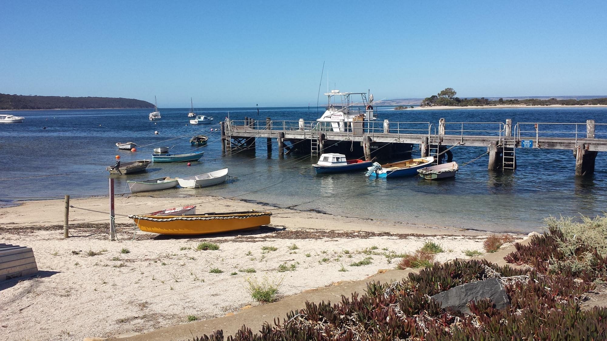 Una marina costera con barcos amarrados en un muelle, playa arenosa y vegetación escasa.