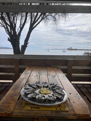 A wooden picnic table with a plate of oysters and lemon slices, overlooking a calm body of water with boats. - Riverside Nature Retreat • Sea Views of Kangaroo Island (American River)