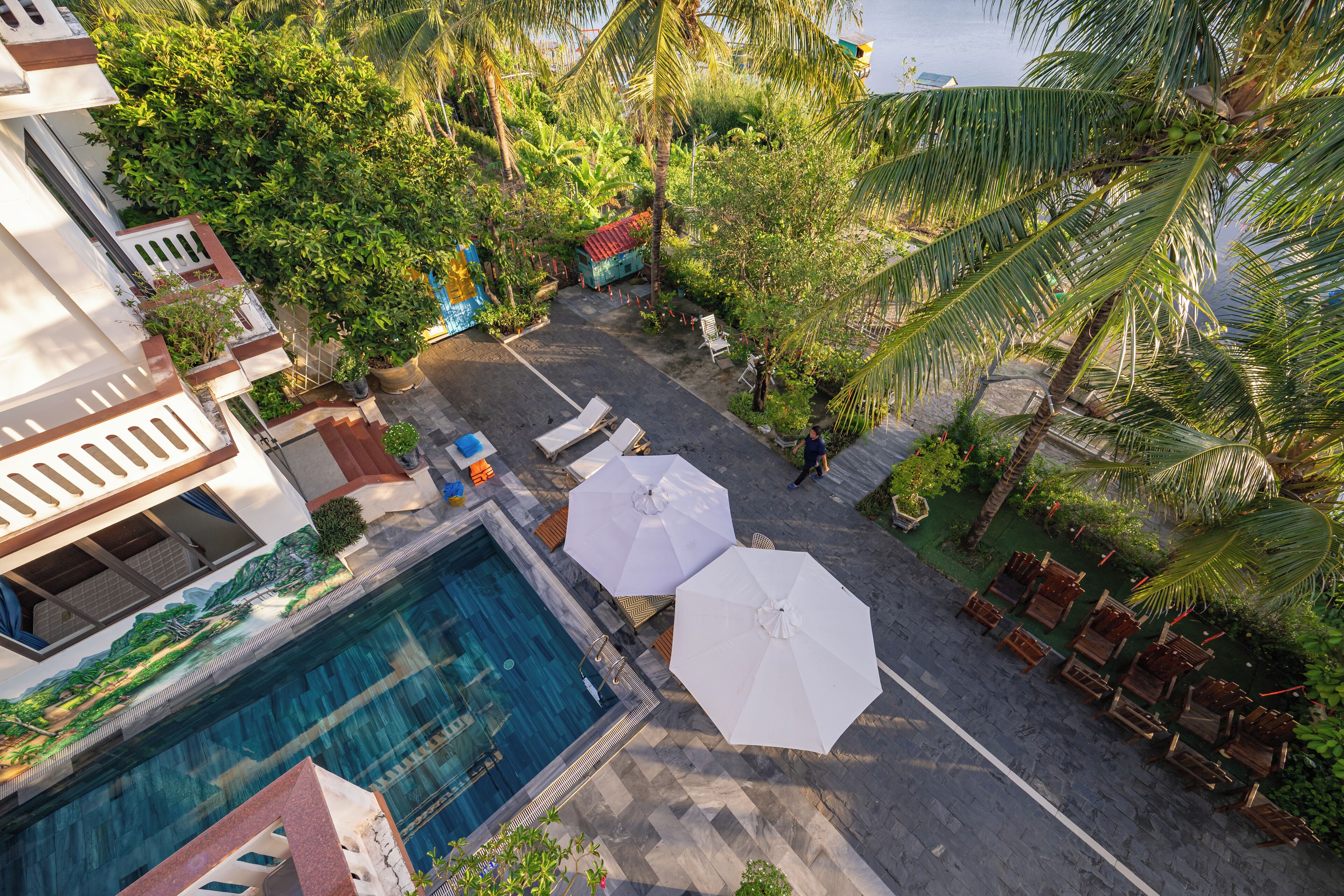 A rooftop pool area with lounge chairs and umbrellas, surrounded by lush greenery and palm trees.