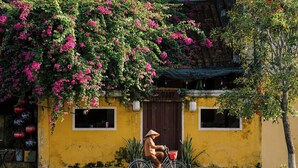 A person riding a bicycle with a basket in front of a yellow building with a wooden door and windows. - CocoPalm Hoi An River Beach By JM Villas (Hoi An)