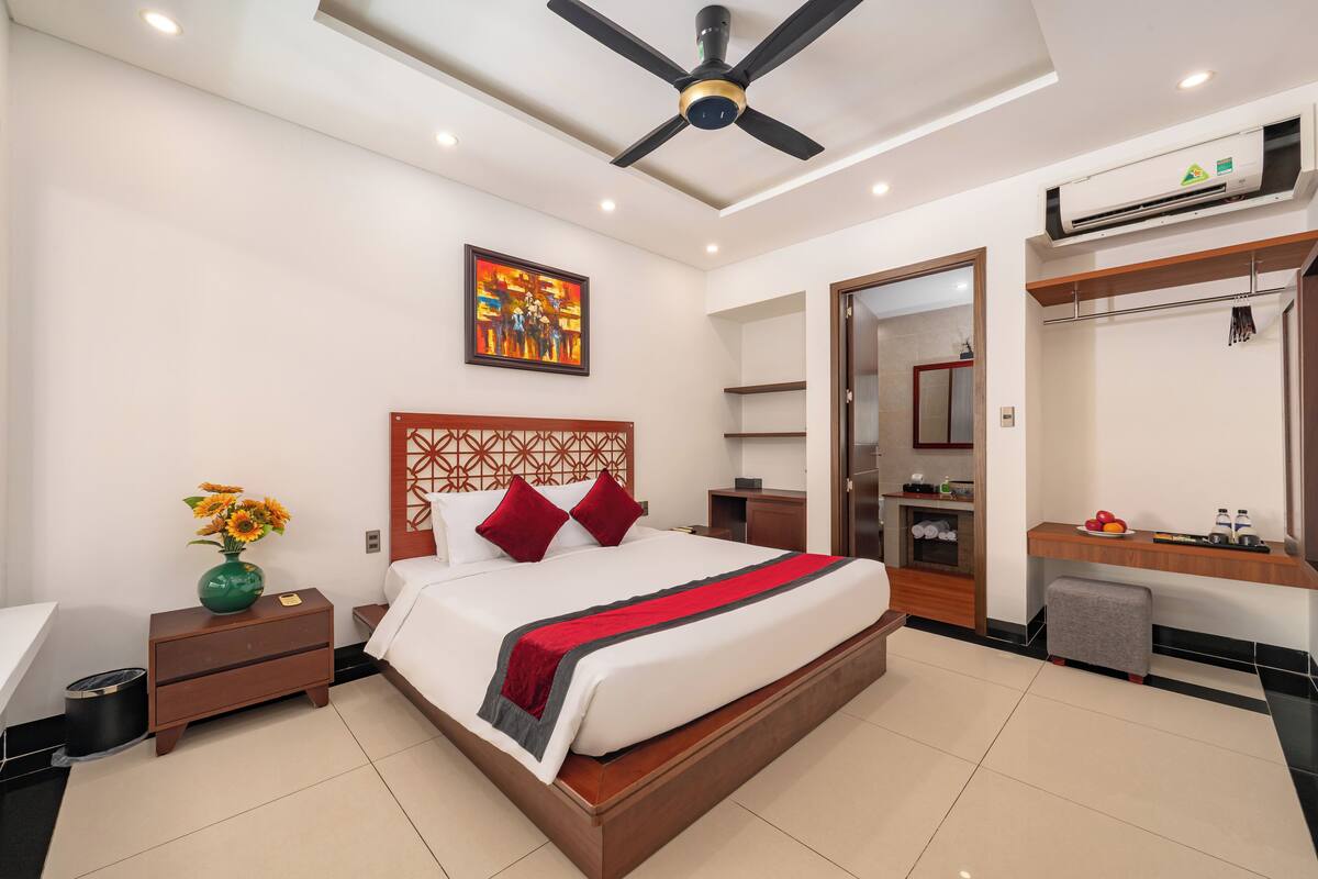 Una habitación de hotel moderna con una cama grande, un ventilador de techo, aire acondicionado de pared, un tocador con espejo y un pequeño taburete.