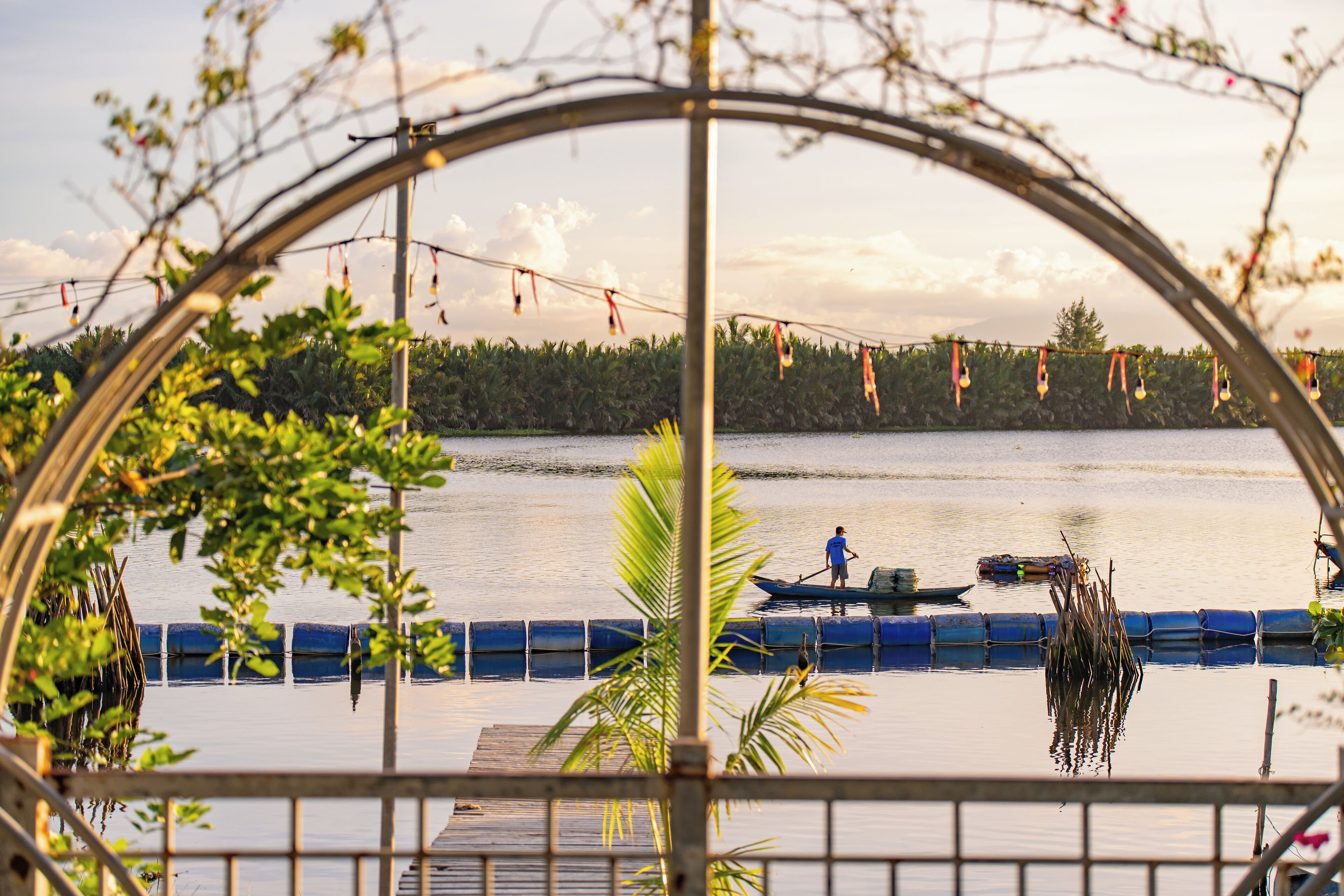 A view through a metal archway of a calm lake with a dock and a person rowing a boat.