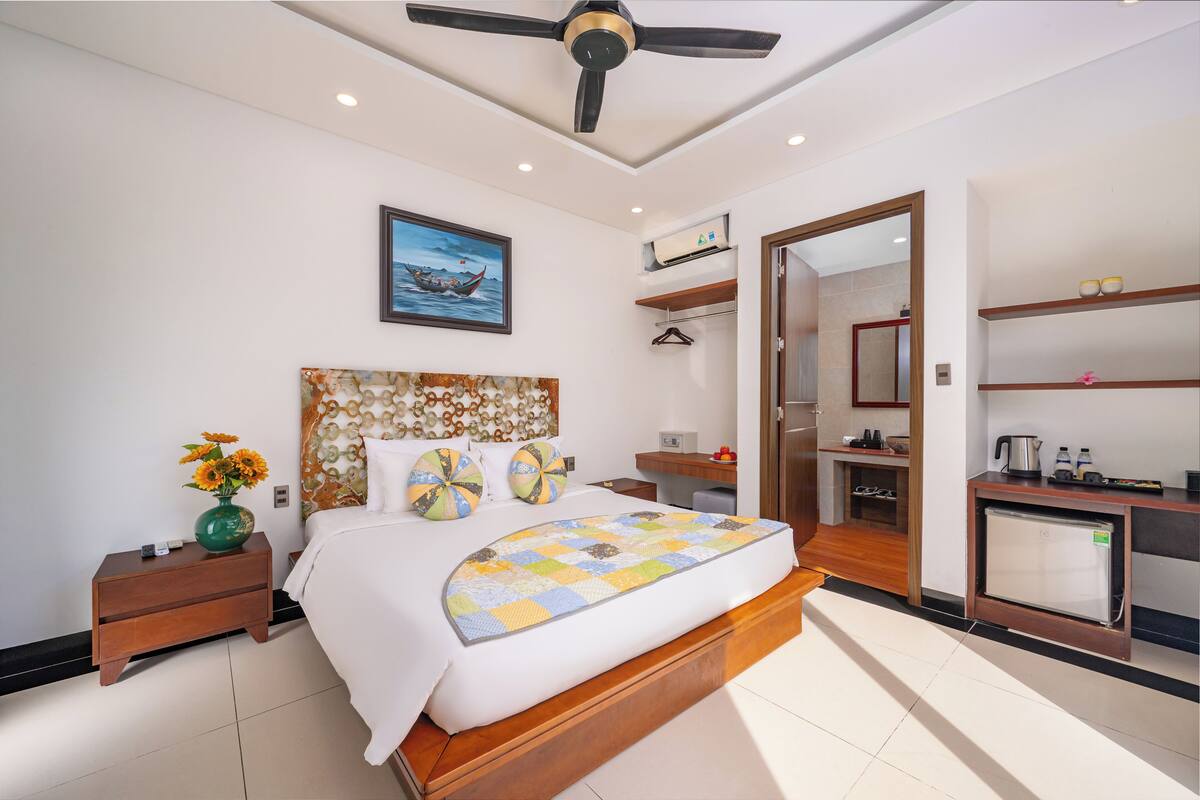 Un dormitorio moderno con una cama grande, una mesita de noche con un jarrón de flores, un ventilador de techo, un cuadro en la pared y una pequeña zona de kitchenette.