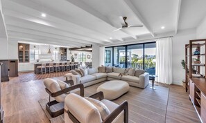 A spacious living area with a large L-shaped sofa, a dining table, and a kitchen in the background. - New Canal Front Beach House on AMI! Gorgeous Pool! (Anna Maria)