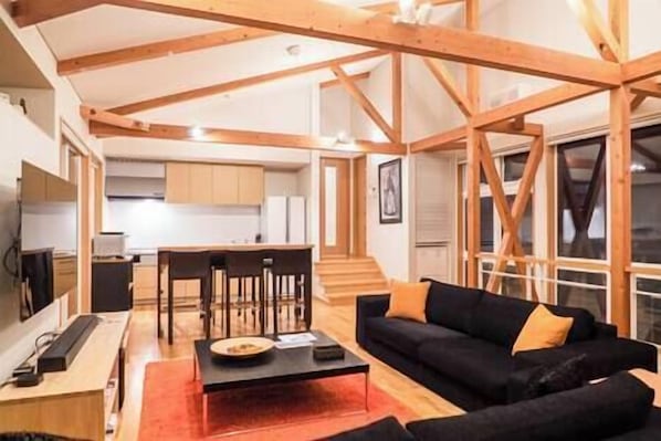 A modern living room with a black sofa, wooden beams, a dining area with chairs, and a kitchen in the background.