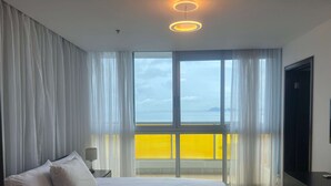 Een slaapkamer met een bed, nachtkastje, lamp, televisie en uitzicht op zee door grote ramen.
