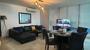 Een moderne woonkamer met een modulaire bank, een eettafel met stoelen, een tv waarop een film wordt afgespeeld, en een plafondlamp.