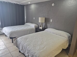 A hotel room with two beds, a nightstand with a lamp, a wall-mounted heater, and a plain grey textured wall. - Budget Inn (Kissimmee)