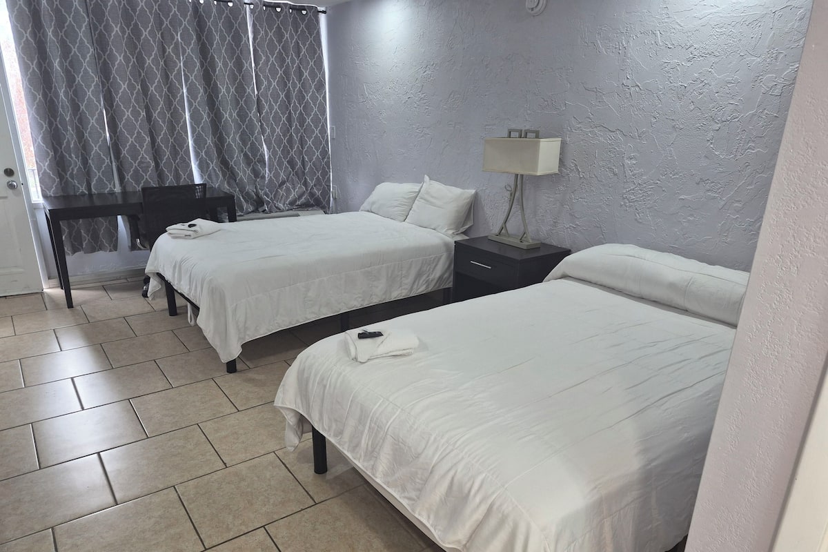 Hotellihuone, jossa on kaksi sänkyä, työpöytä, lamppu ja ikkuna, jossa on verhot.