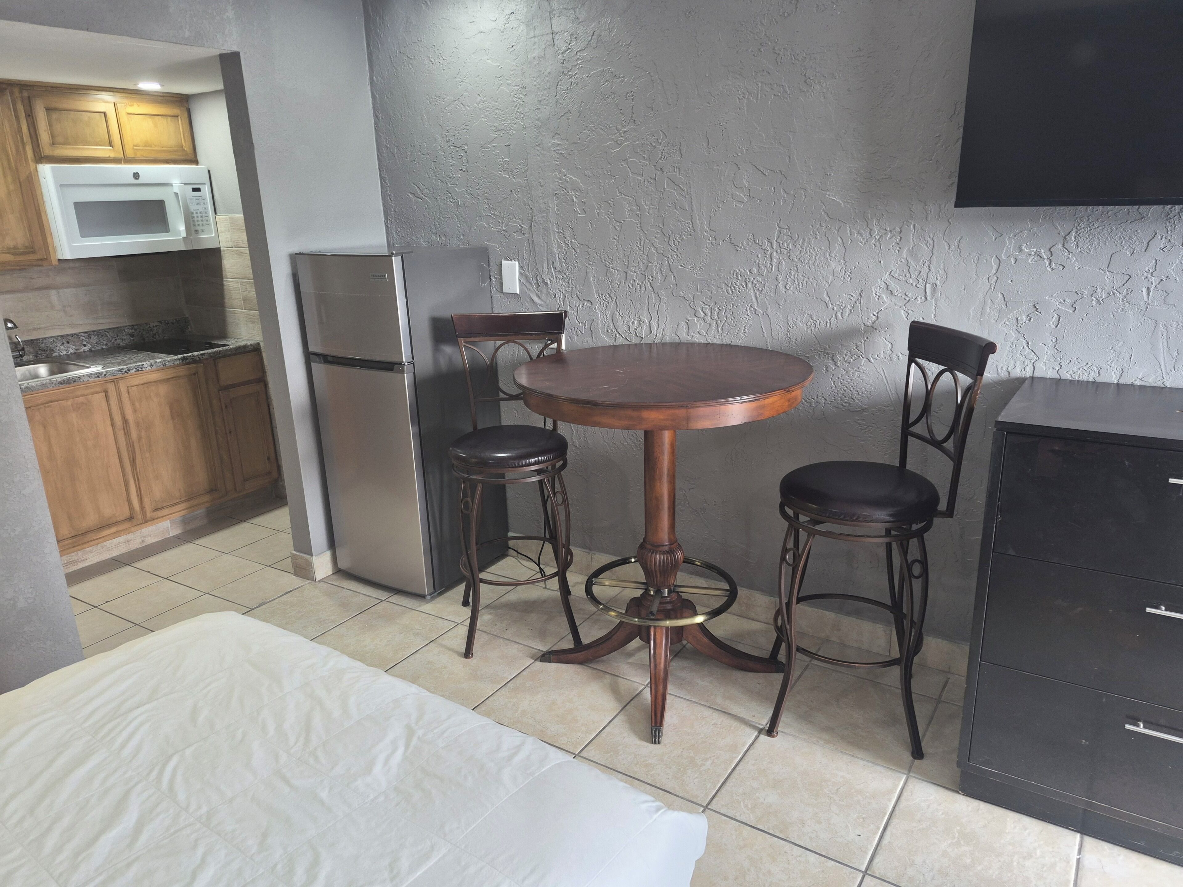 Cocina compacta con microondas, refrigerador y armarios de madera. Hay una mesa redonda con dos sillas y un mueble negro.