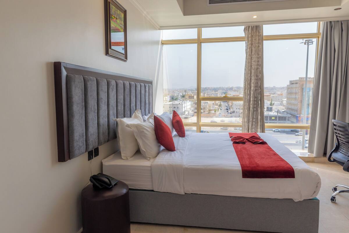 A hotel room with a large window, a bed with white and red bedding, a grey headboard, and a small bedside table with a lamp.
