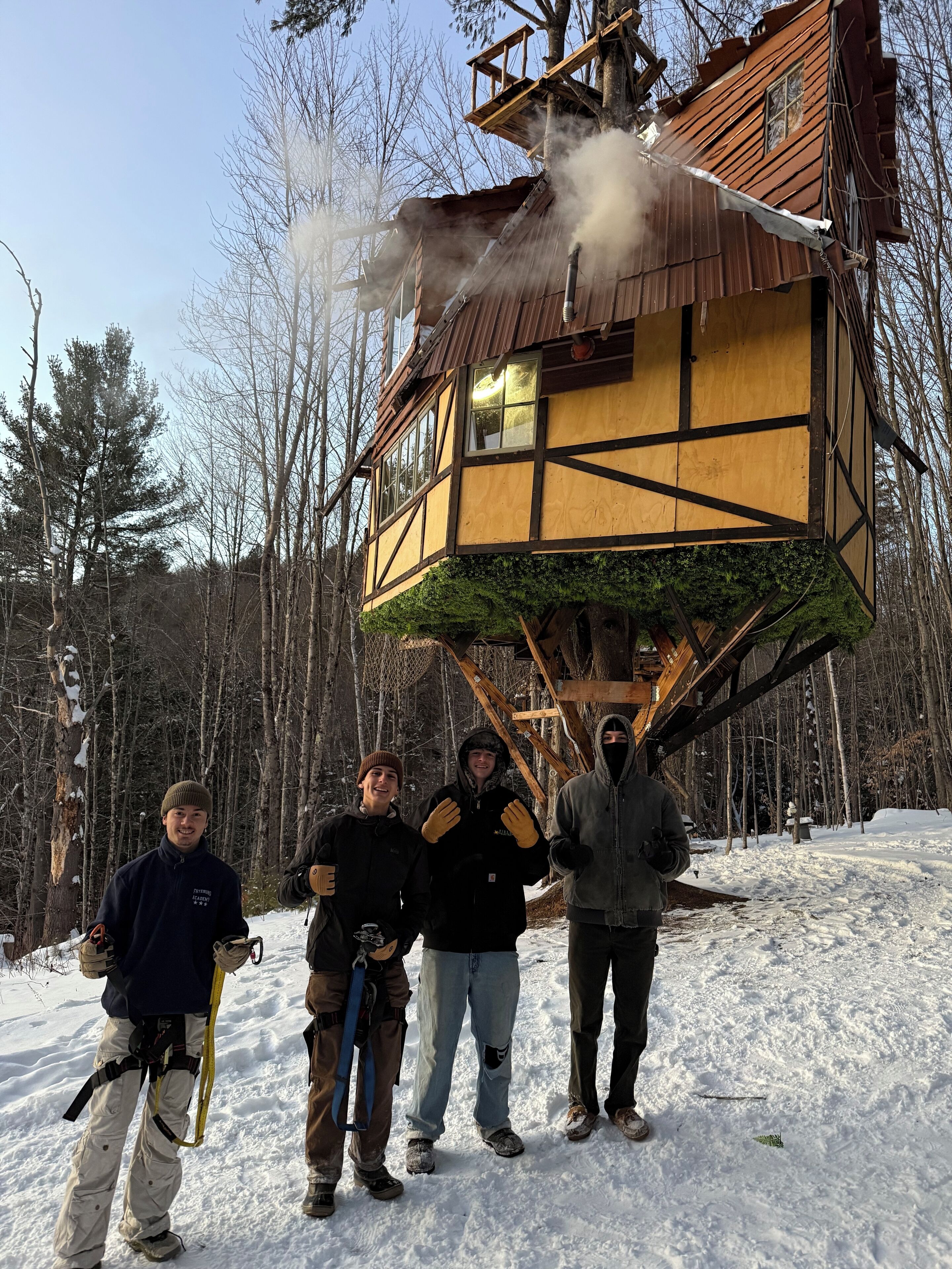 Une cabane en bois, dotée d’un toit à forte pente et d’une cheminée d’où s’échappe de la fumée, est entourée d’une forêt enneigée.