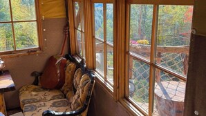 A room with a wooden bench, a guitar, and large windows offering a view of a forested area. - Charming TREEHOUSE in remote location (Braintree)
