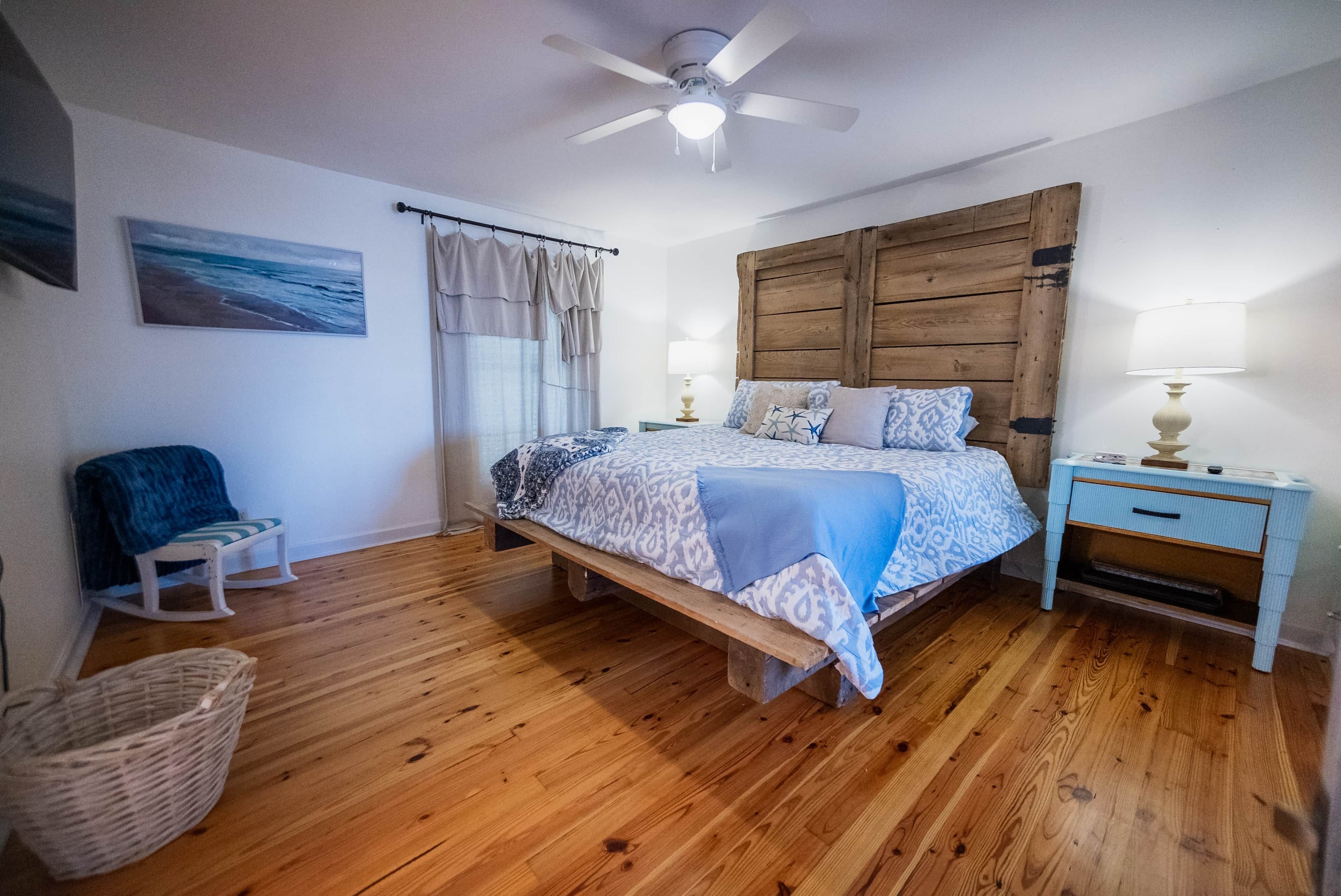 Un dormitorio con cama de madera, mesita de noche, silla, ventilador de techo montado en la pared y un cuadro con motivos playeros.