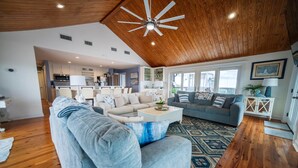 A spacious living room with a high ceiling, wooden flooring, and a large window providing natural light.