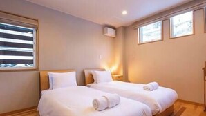 Quarto de hotel com duas camas, janela com persianas, ar condicionado fixado na parede e mesa de cabeceira com luminária.