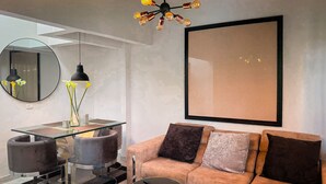 Una sala moderna con una mesa de comedor de vidrio, un sofá con varios cojines, una obra de arte grande enmarcada en la pared y una lámpara de techo original.