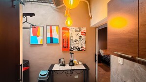 Habitación compacta con kitchenette, una mesita con hervidor de agua, un cesto de basura y una cama al fondo.