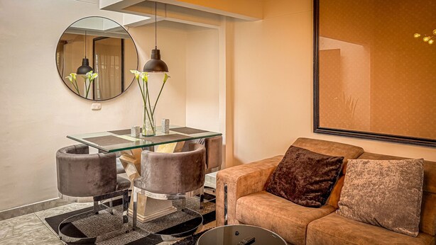 Una sala moderna con una mesa de comedor de vidrio, dos sillones acolchados, un sofá con un cojín marrón, un espejo redondo y una lámpara colgante.