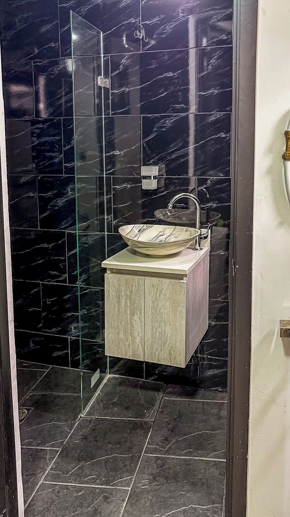 A bathroom with a glass-enclosed shower, a marble-tiled wall, and a wooden vanity with a bowl sink.