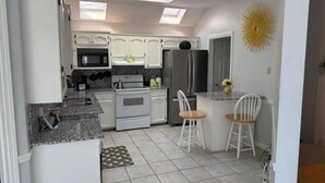 Une cuisine compacte avec des meubles blancs, un four encastré, un four à micro-ondes et un réfrigérateur. On y trouve un petit îlot central doté d’un évier et de deux chaises.