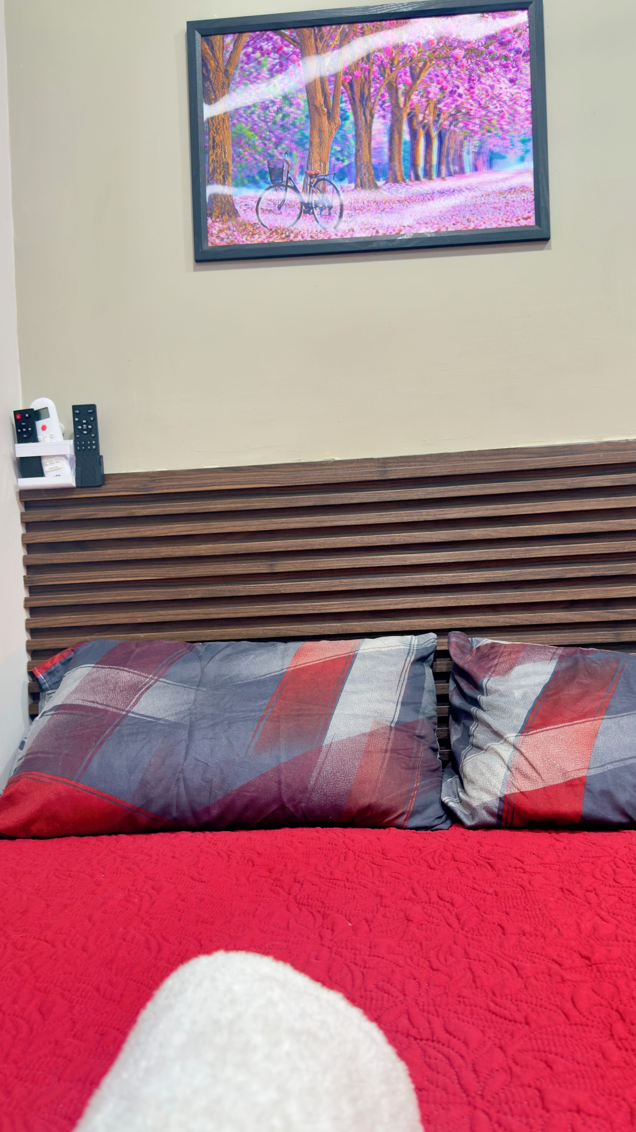 Una cama con una colcha roja y almohadas escocesas, un cabecero de madera y un cuadro con una bicicleta en un parque.