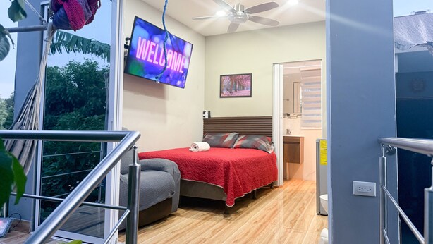 Un dormitorio moderno con una colcha roja, un televisor que muestra 'WELCOME', un ventilador de techo y vistas al exterior.