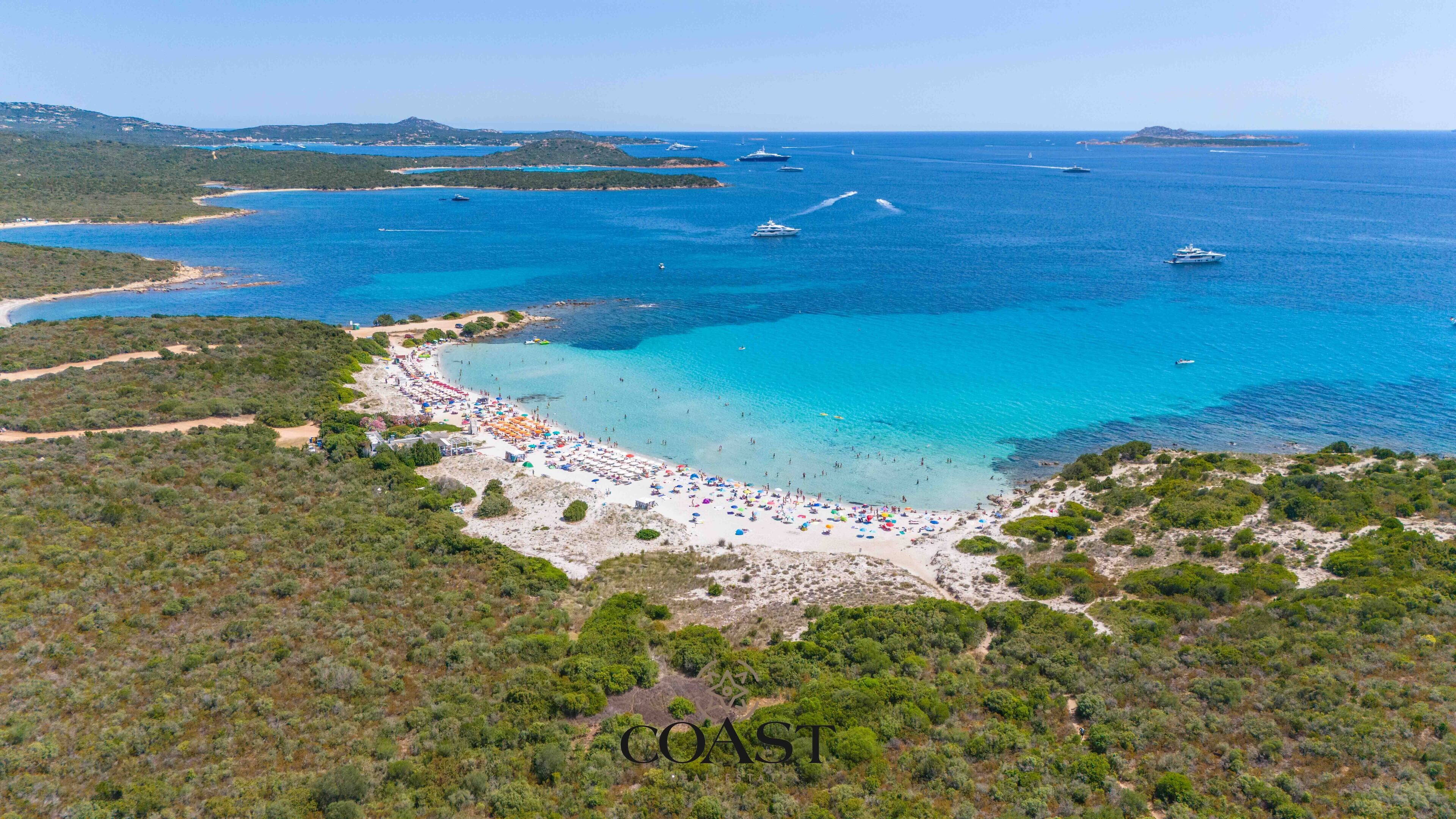Vista aérea de una zona costera con playa, barcos y aguas azules cristalinas.